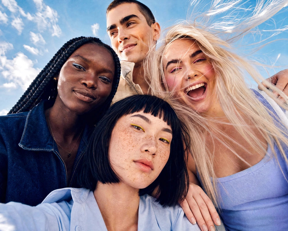 Four young adults taking a close-up selfie outdoors under a bright blue sky with clouds. Their hair is blowing in the wind, and they are wearing casual light blue and navy tops.